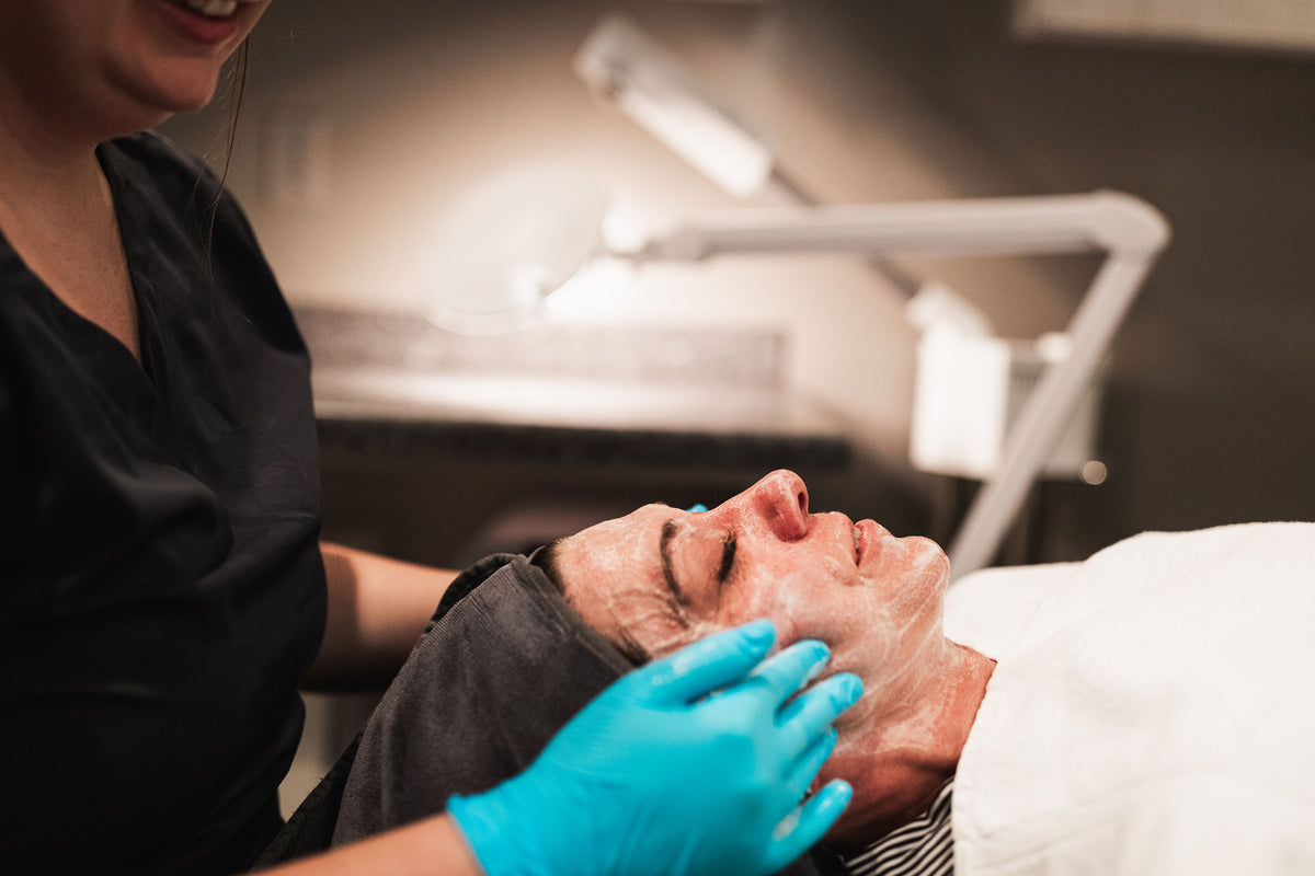 Smiling clinician in black scrubs applying a creamy facial mask to a reclining patient at Element Dermatology