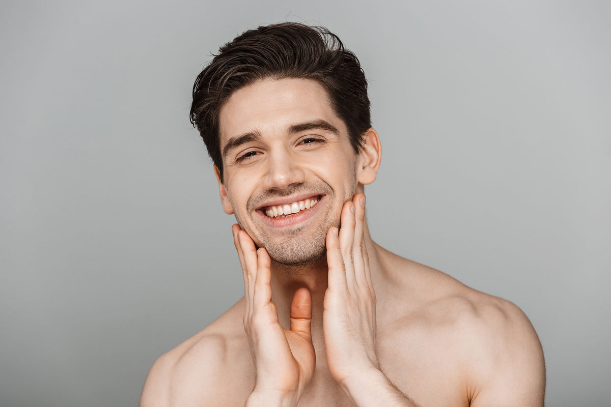 Smiling man touching his smooth jaw and cheeks with both hands against a grey background