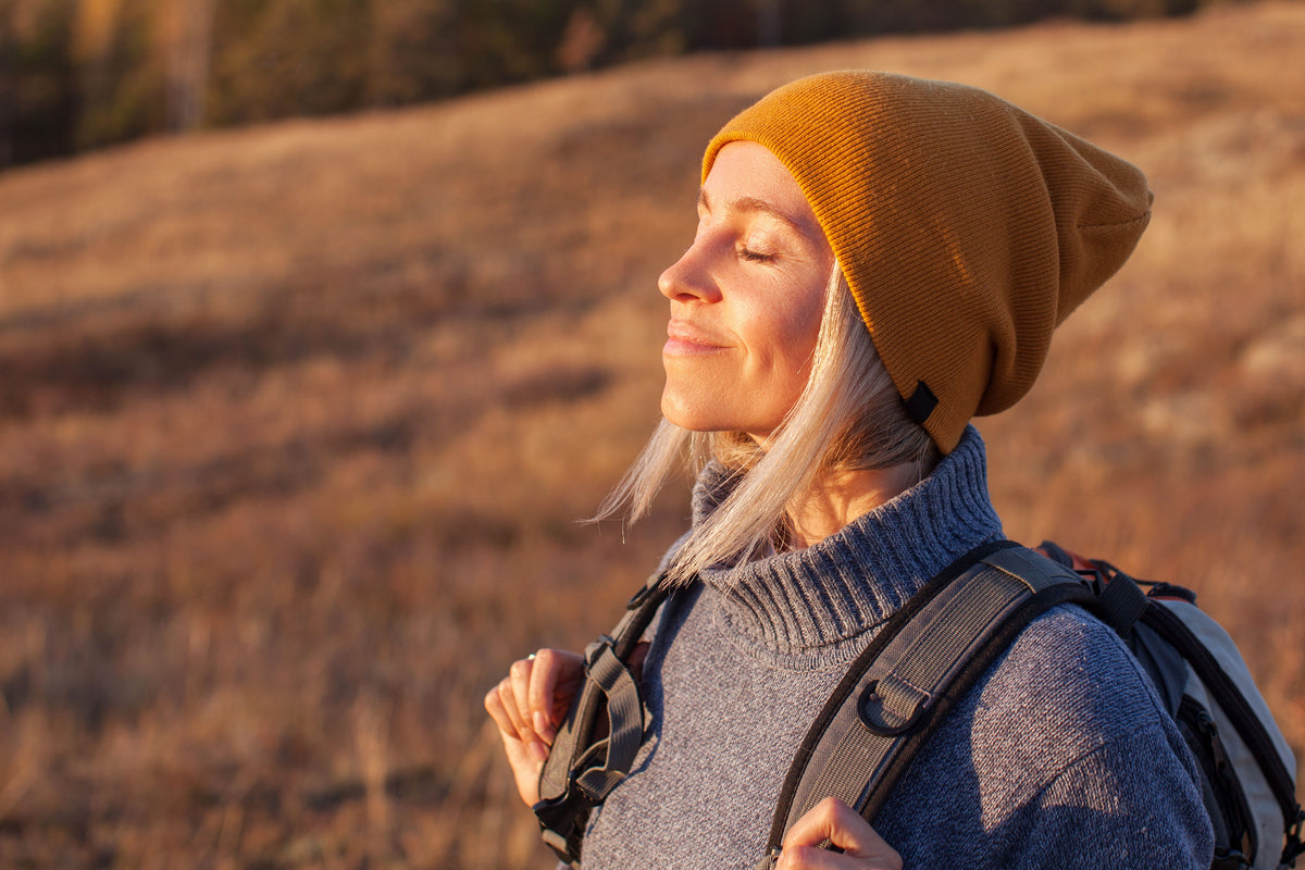 Woman smiling outdoors in warm golden light, wearing a mustard beanie with eyes closed