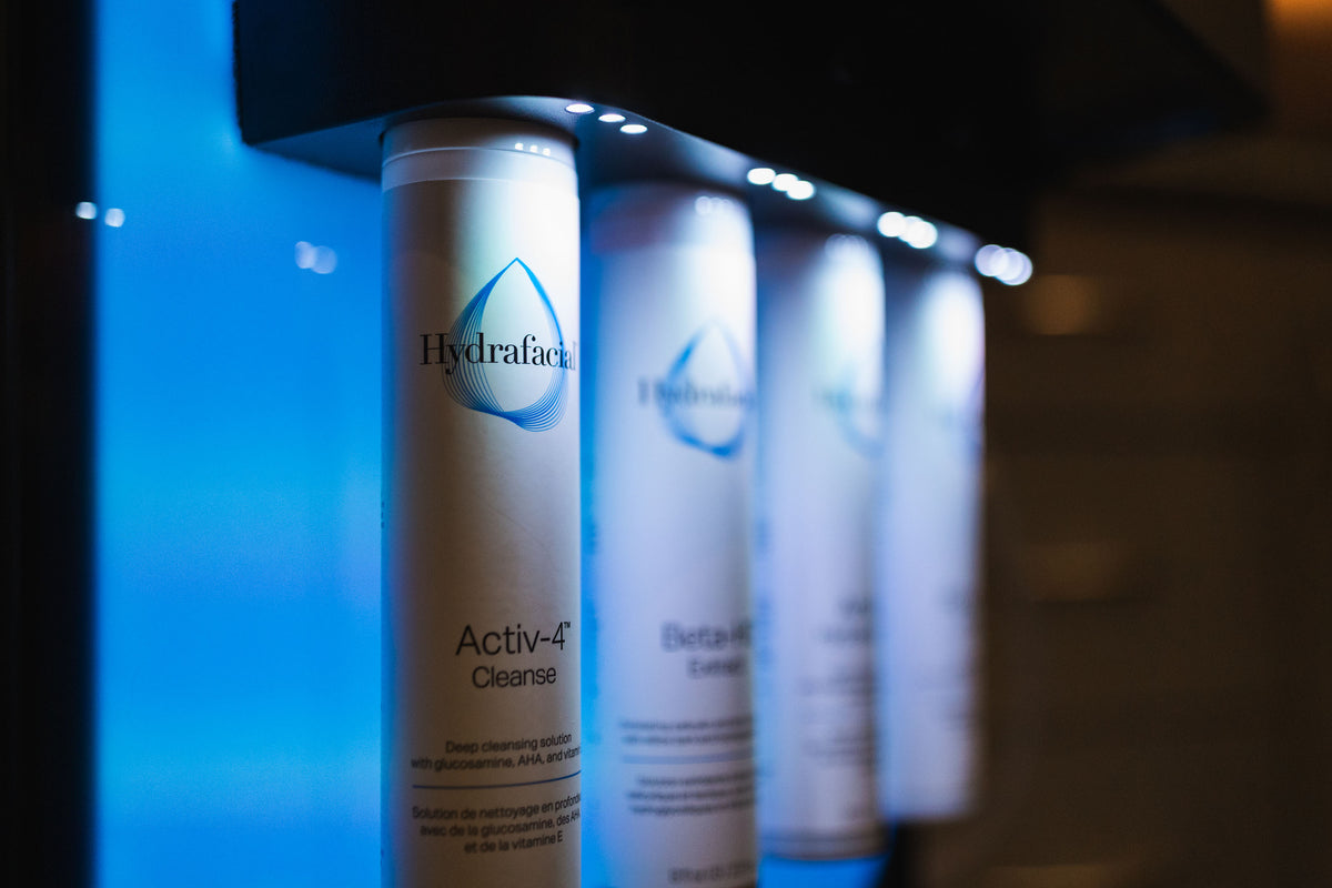 Row of backlit HydraFacial solution bottles, including Activ-4 Cleanse, glowing in blue light