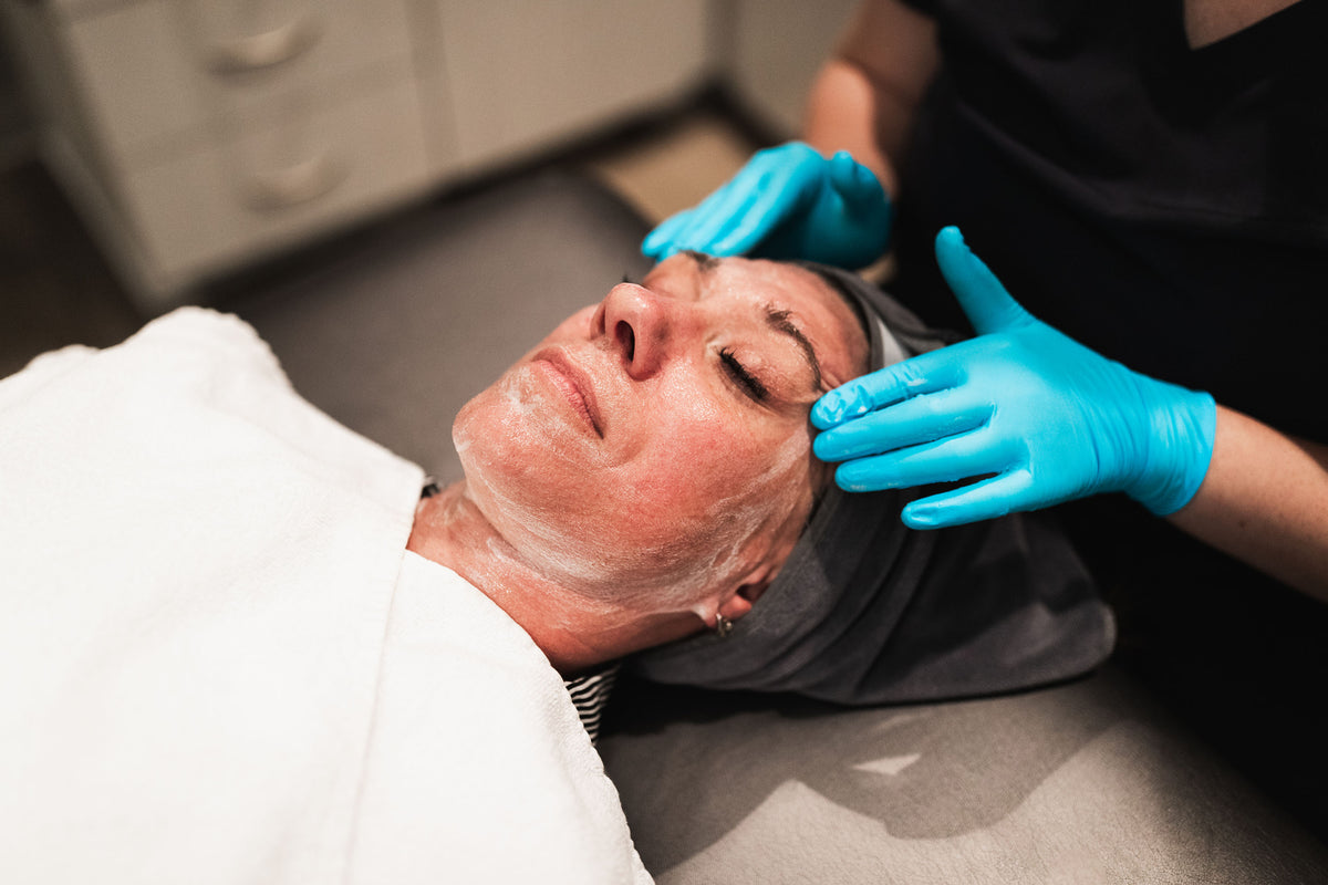 Gloved clinician applying a white exfoliating mask to a reclining patient's face at Element Dermatology