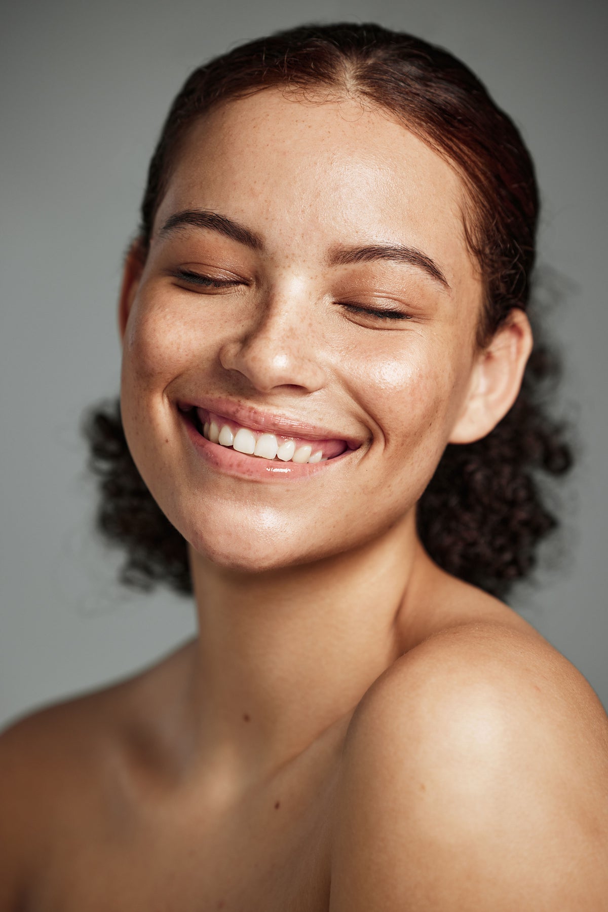 Young woman with freckles and curly hair smiling with eyes closed against a grey background