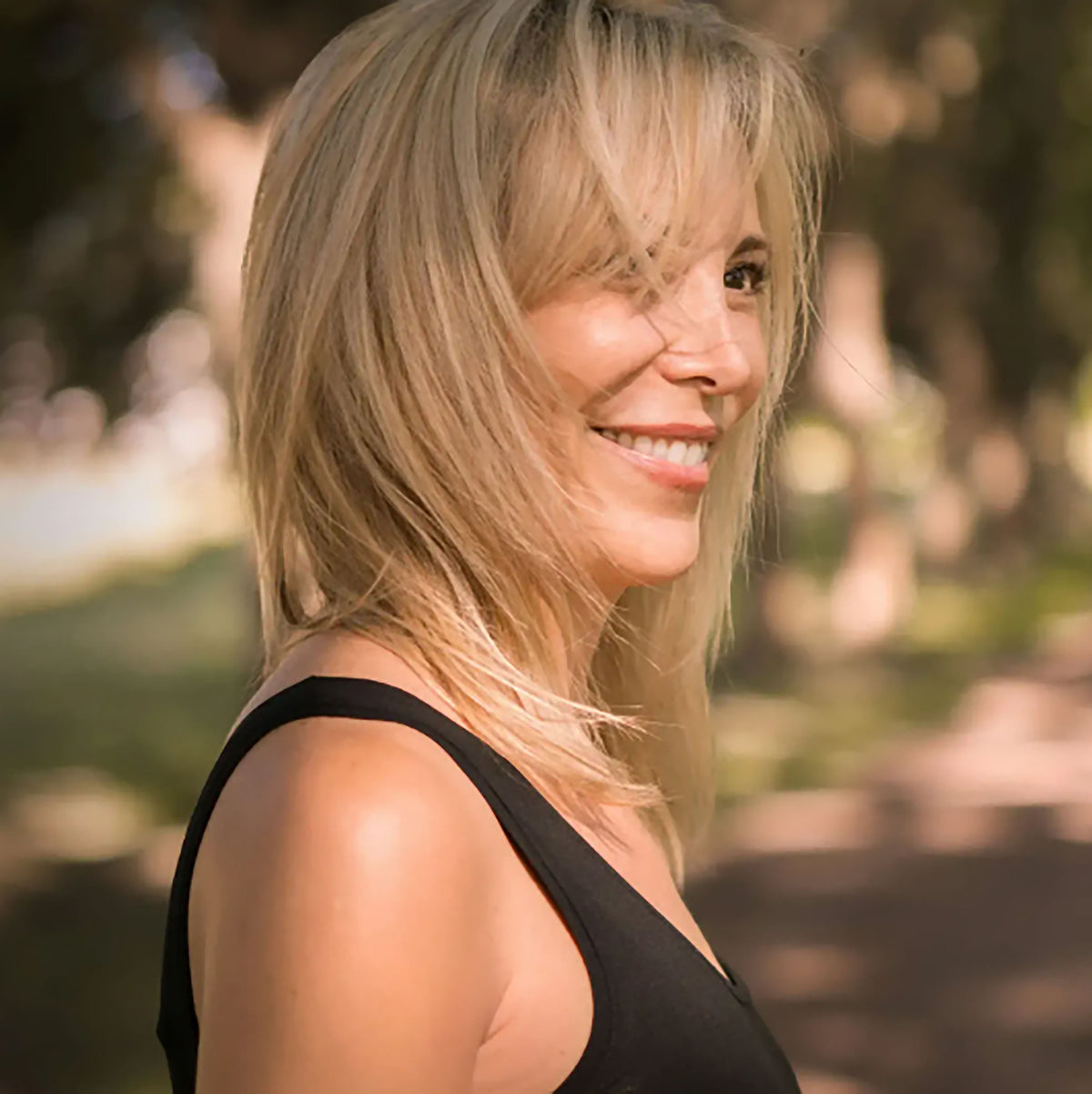 Smiling woman with blonde hair outdoors in natural light, wearing a black sleeveless top