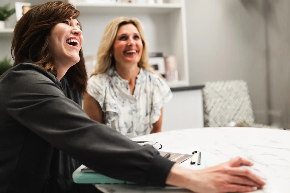 Two women laughing together at a table in a bright, modern consultation room